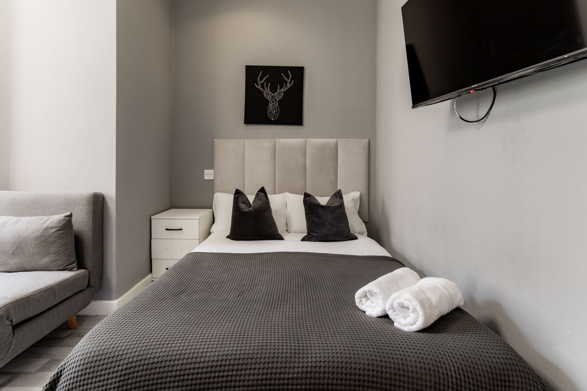 A modern hotel room with a large bed, a flat-screen TV mounted on the wall, a small bedside table, and a grey sofa.