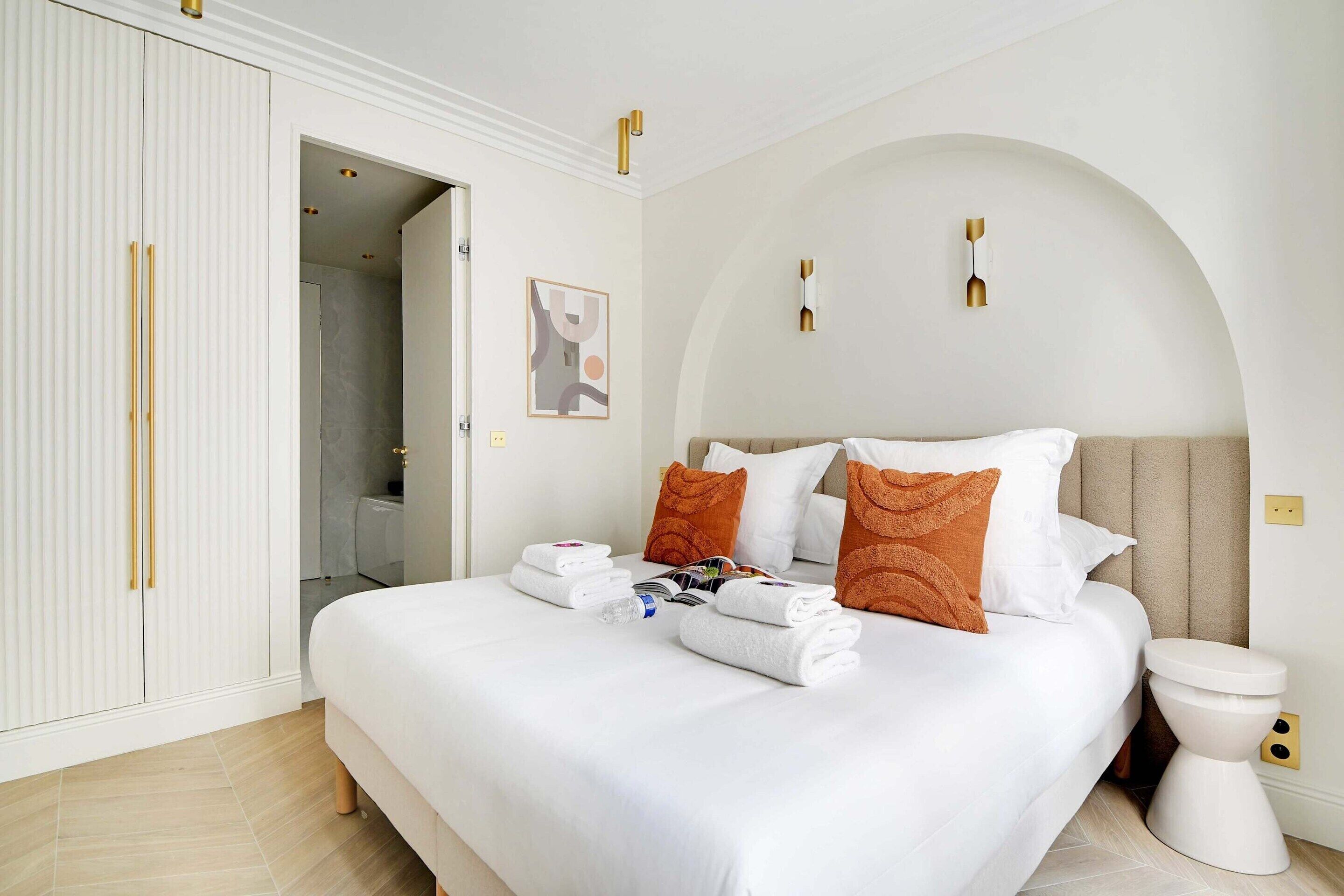 A neatly made bed with white linens and orange pillows, a white wardrobe with gold handles, and a modern bathroom with a white toilet.
