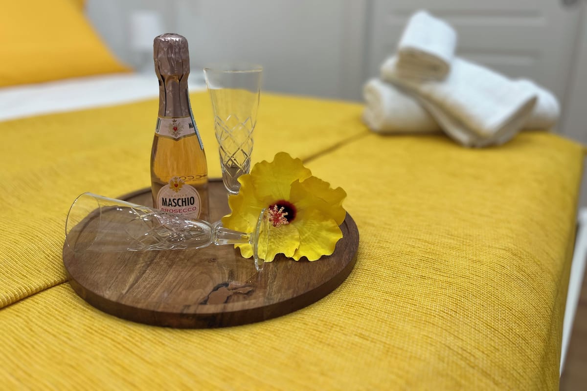 A wooden tray with a bottle of Maschio prosecco, a glass, and a yellow flower on a yellow bedspread.