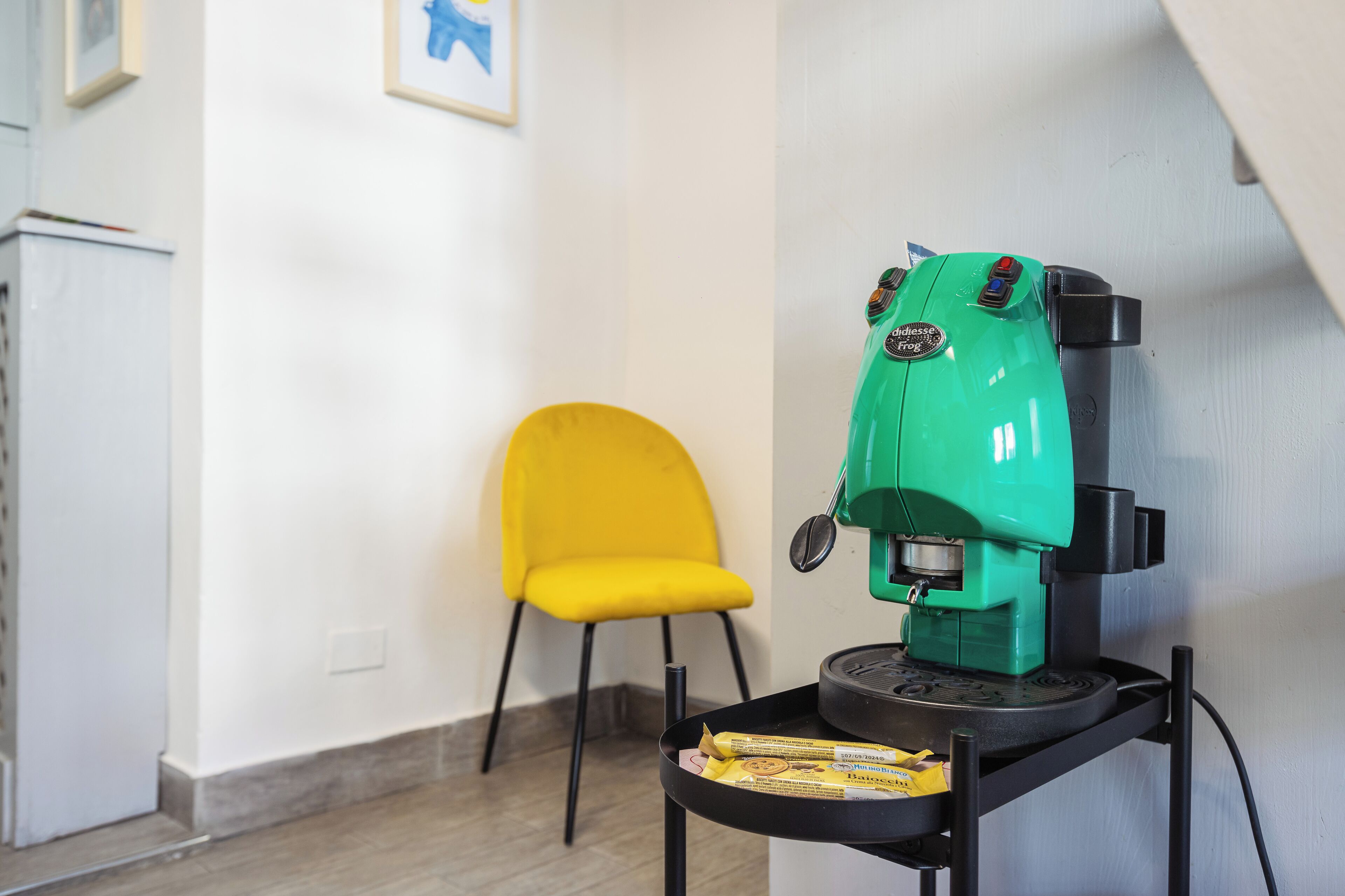 A room with a green coffee machine on a black stand, a yellow chair, and a white wall with a framed picture.