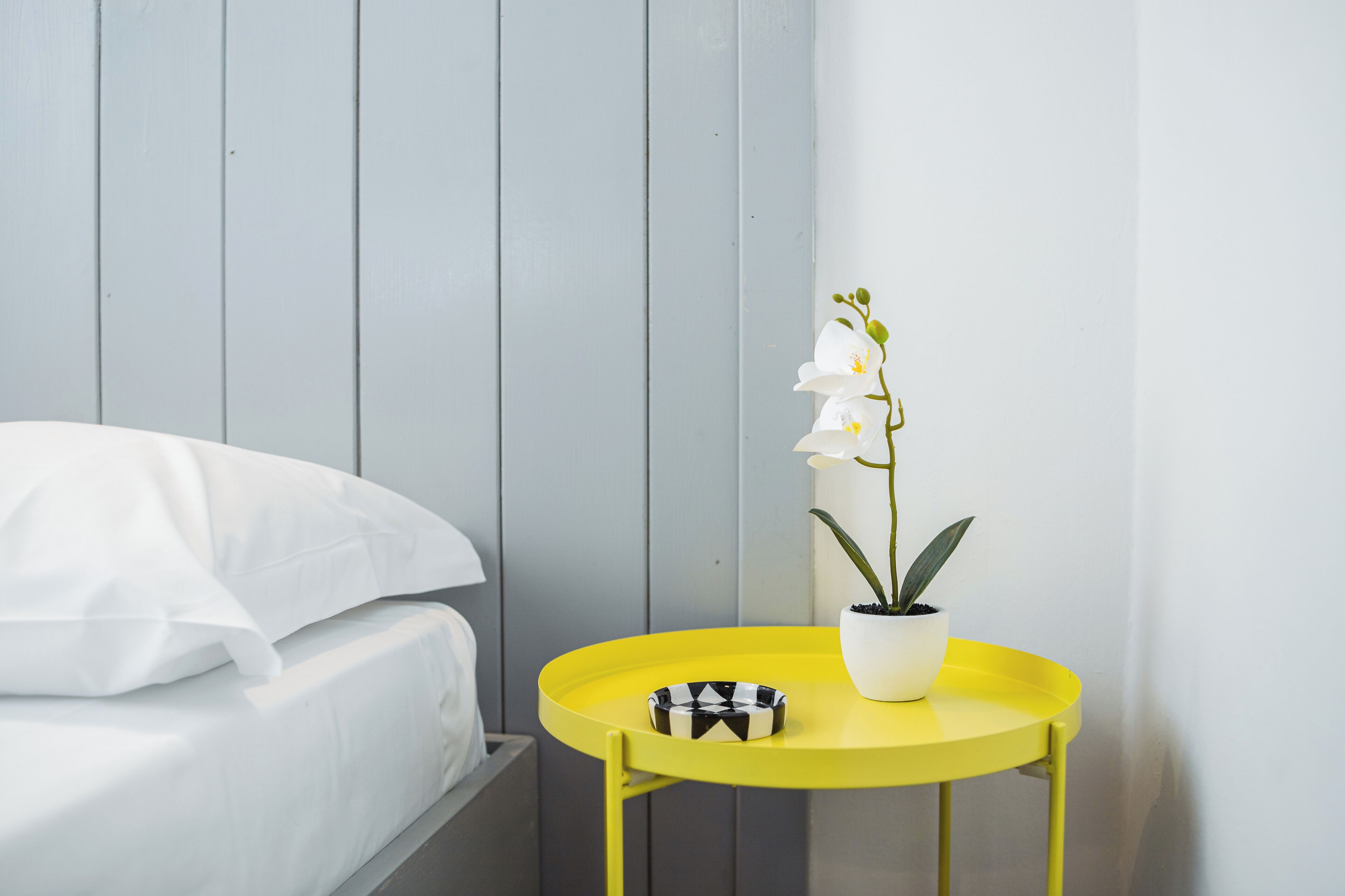 A bed with white bedding, a yellow side table with a potted orchid, and a black and white bowl.