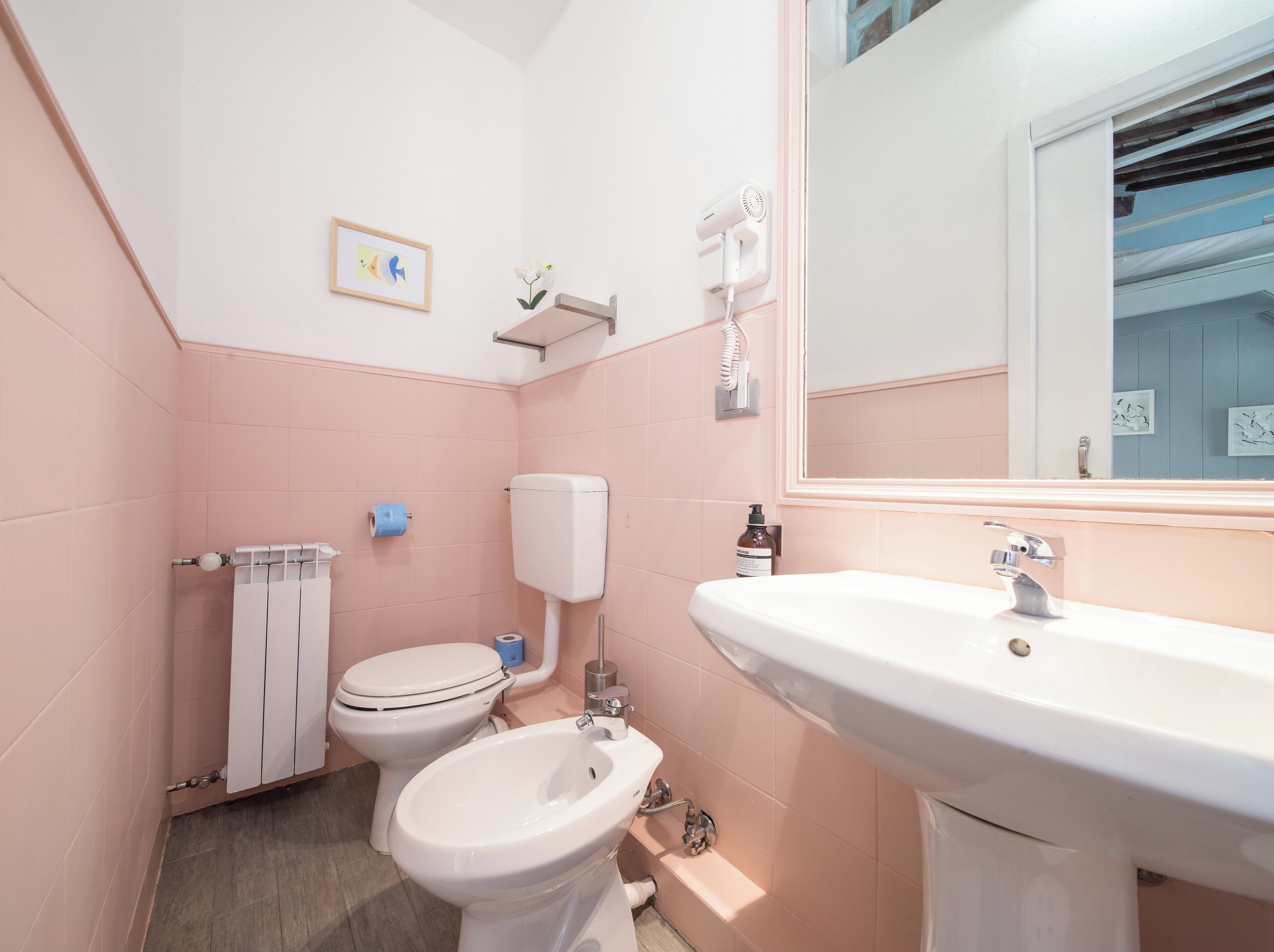 Une salle de bain avec des murs roses, un lavabo blanc, des toilettes et un bidet. Un tableau encadré est accroché au mur, et un sèche-cheveux est fixé au mur.