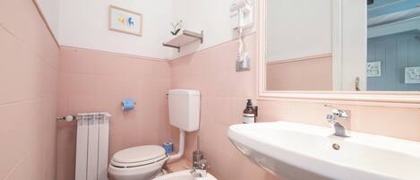 Une salle de bain avec des murs roses, un lavabo blanc, des toilettes et un bidet. Un tableau encadré est accroché au mur, et un sèche-cheveux est fixé au mur.