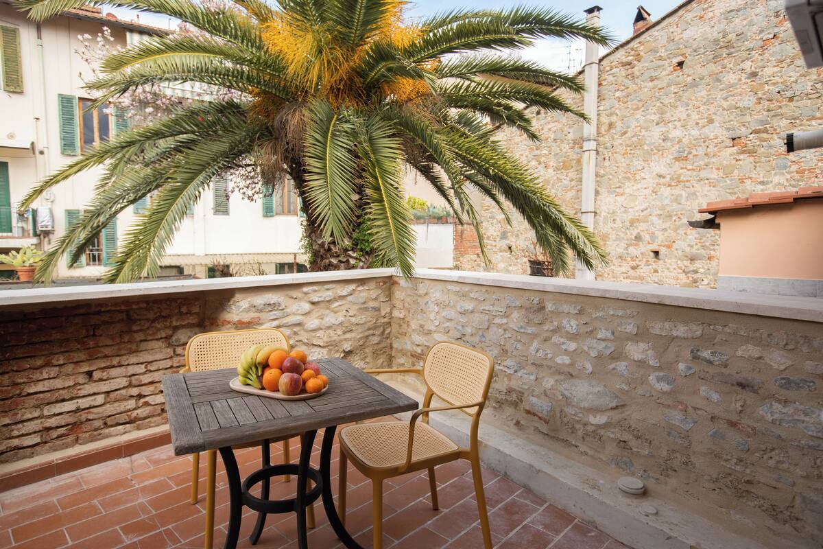 A balcony with a table, chairs, a fruit bowl, and a palm tree.