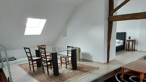 Ein geräumiger Loft mit Essbereich, der einen Glastisch und Holzstühle, ein sichtbares Dachfenster und eine hohe Decke mit freiliegenden Balken zeigt.