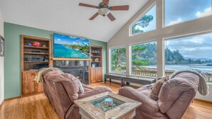 A living room with a large TV, wooden shelves, a fireplace, and a view of the ocean.