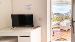 Ett rum med en platt-TV på ett vitt skrivbord, ett litet skåp under skrivbordet och en dörr som leder ut till en uteplats med en vit stol och ett bord.