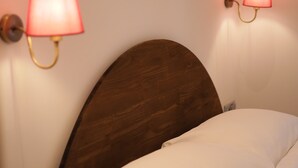 Cama bem arrumada com lençóis brancos e cabeceira de madeira. Duas luminárias fixadas na parede com cúpulas vermelhas em ambos os lados da cama.