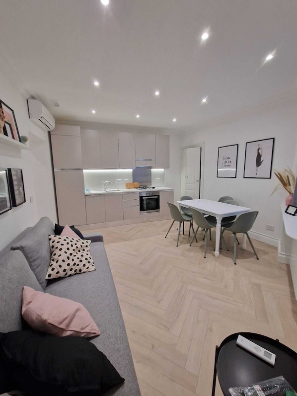 Un apartamento moderno con un sofá gris, una mesa de comedor con sillas, una zona de cocina con armarios y un aire acondicionado instalado en la pared.