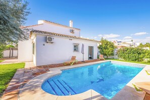 Une piscine avec une terrasse carrelée, un bâtiment blanc avec un toit recouvert de tuiles et une pelouse verte.