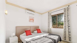 A bedroom with two beds, a nightstand, an air conditioner, a window with a view, and a decorative wall piece. - Villa Bart (Estepona)