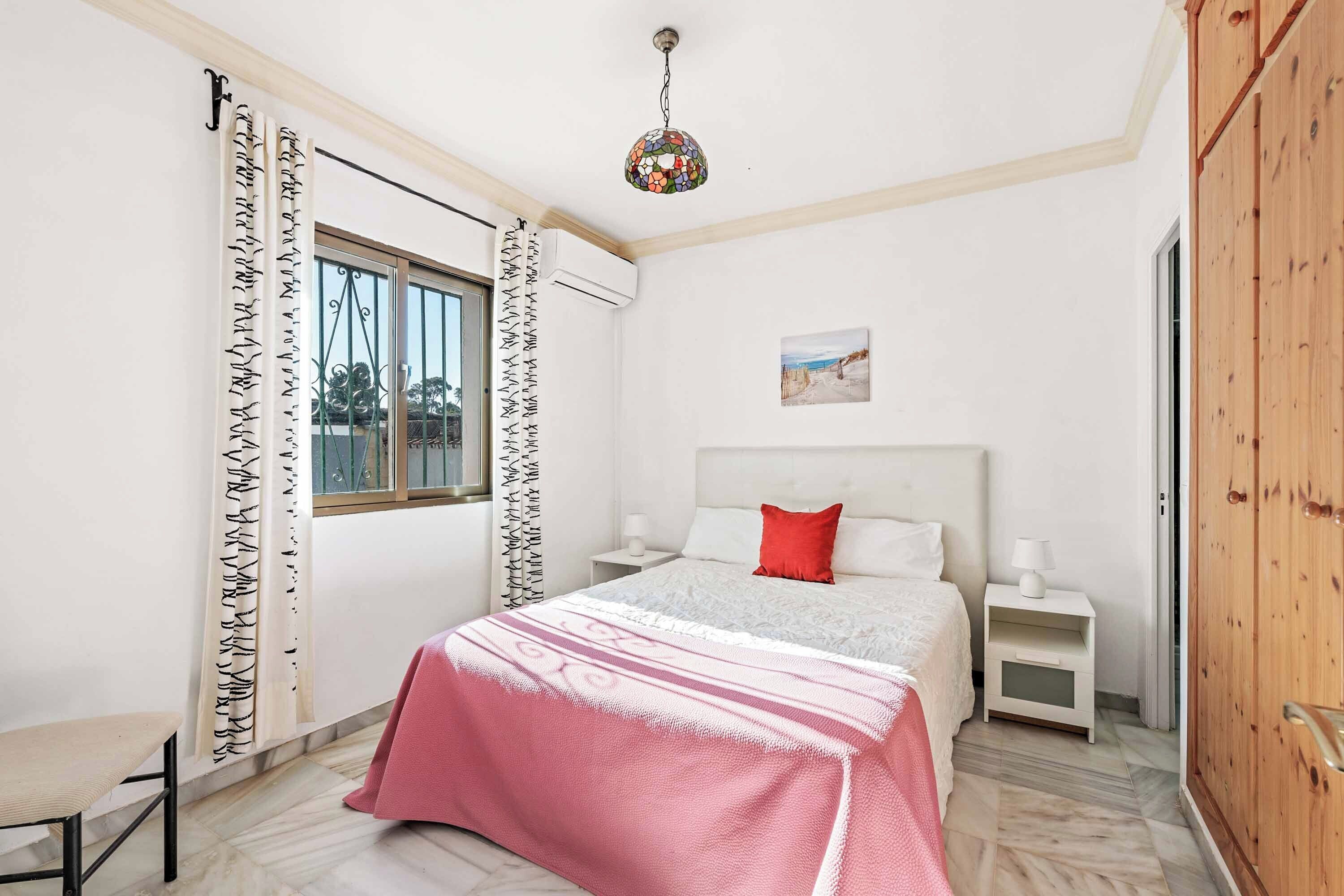 Un dormitorio con cama, mesitas de noche, una silla, un ventanal con cortinas y un cuadro en la pared.