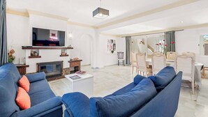 A modern living room with a flat-screen TV, blue sofas, a fireplace, and a dining area with chairs and a table. - Villa Bart (Estepona)