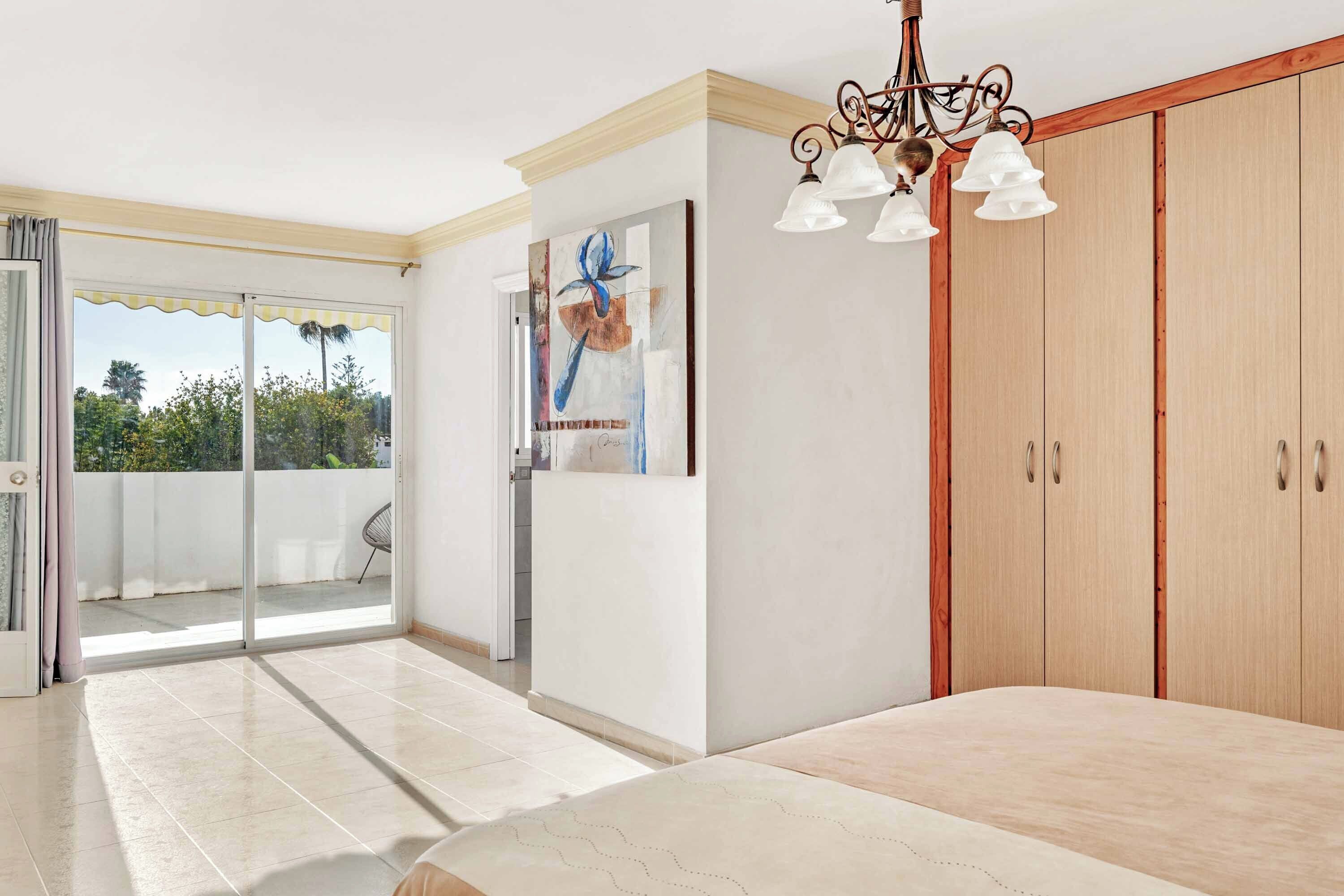 A bedroom with a bed, a chandelier, a wardrobe, and a sliding glass door leading to a balcony.