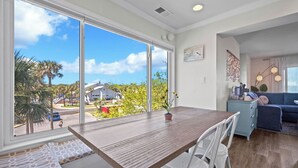 A dining area with a wooden table and white chairs, large windows with a view of a residential neighborhood, and a blue sofa in the background. - Ocean View Condo, Golf Cart Included, Private Patio, Newly Redecorated - 607 Newhaven (Fripp Island)