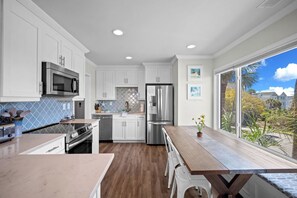 A modern kitchen with white cabinets, stainless steel appliances, a wooden dining table, and a view of a residential area. - Ocean View Condo, Golf Cart Included, Private Patio, Newly Redecorated - 607 Newhaven (Fripp Island)
