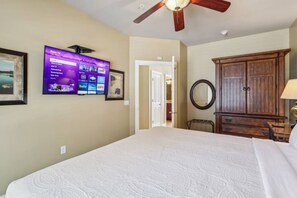 Quarto de hotel com uma cama grande, um ventilador de teto, uma TV fixada na parede, um guarda-roupa de madeira e um espelho redondo.