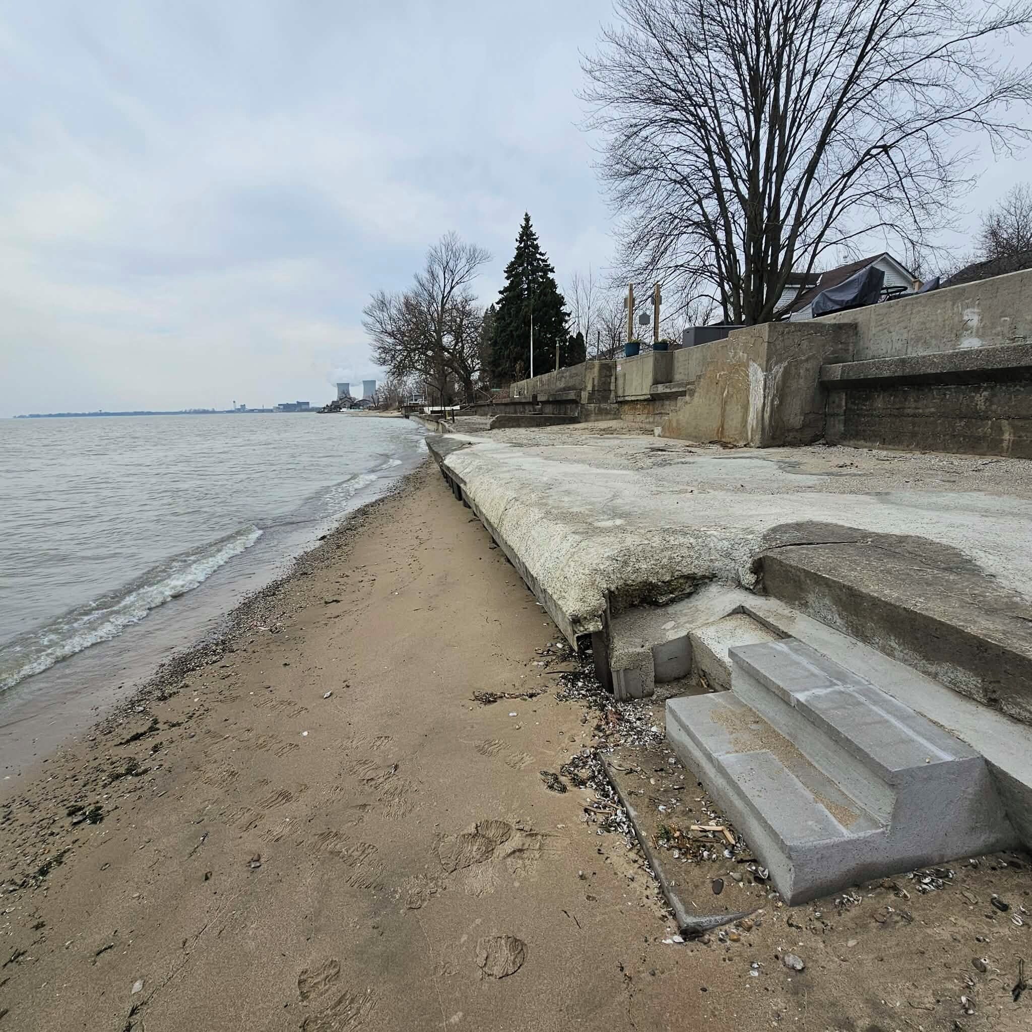 A concrete waterfront promenade with steps leading to a pier, a sandy beach, and leafless trees.