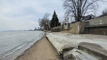 A concrete waterfront promenade with steps leading to a pier, a sandy beach, and leafless trees.