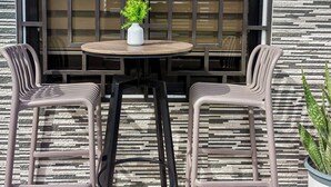 An outdoor seating area with a round table and two chairs, a potted plant, and a window with a geometric pattern.
