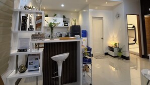 Ein modernes Wohnzimmer mit einem weißen Sofa, einem Couchtisch und einem Essbereich mit weißem Tisch und Stühlen. Es gibt eine Küchenzeile mit Kühlschrank, Mikrowelle und Spüle.