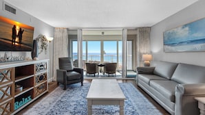 Un salon avec un canapé, une table basse et un téléviseur posé sur un meuble. Il y a un balcon avec vue sur la mer.