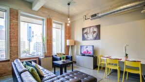 A modern living room with a leather sofa, a dining table with yellow chairs, a TV, and a city view through large windows. - Urban Style 2 BR | 2 Bath | Downtown | Free Valet Parking | 5 (Dallas)