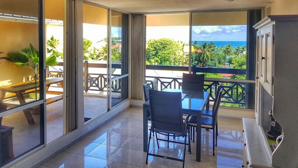 A balcony with a glass door, a table, chairs, and a view of the ocean. - Apartment by the sea (Le Gosier)