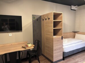 Habitación compacta con escritorio de madera, una silla, una cama y un armario empotrado con ganchos.
