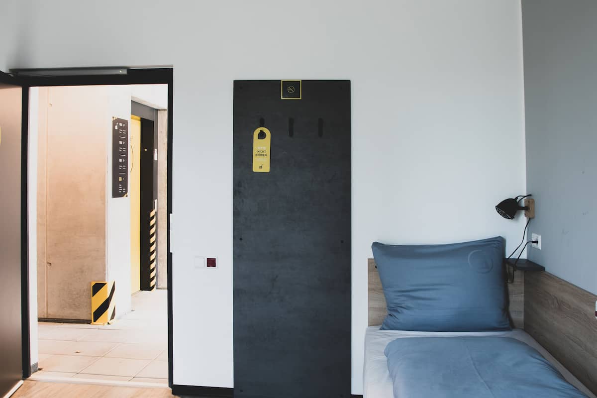 A hotel room with a bed, a bedside lamp, a shelf with towels, and a door with a sign.