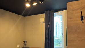 A hotel room with a wooden bed, a small table, a chair, a ceiling light, and a view of the outside through a window. - Das Schlafwerk Köln-Messe (Cologne)