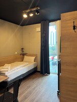 Quarto de hotel com cama de madeira, uma mesinha, uma cadeira, uma luminária no teto e vista para o exterior pela janela.