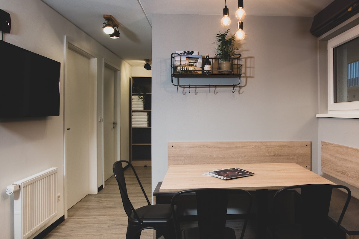 A modern dining area with a wooden table, black chairs, and pendant lighting. A shelf with books and a plant hangs above the table.