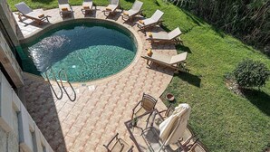 Una piscina circular con sillones de descanso y una mesa, rodeada de un césped cuidado y un patio de ladrillo.