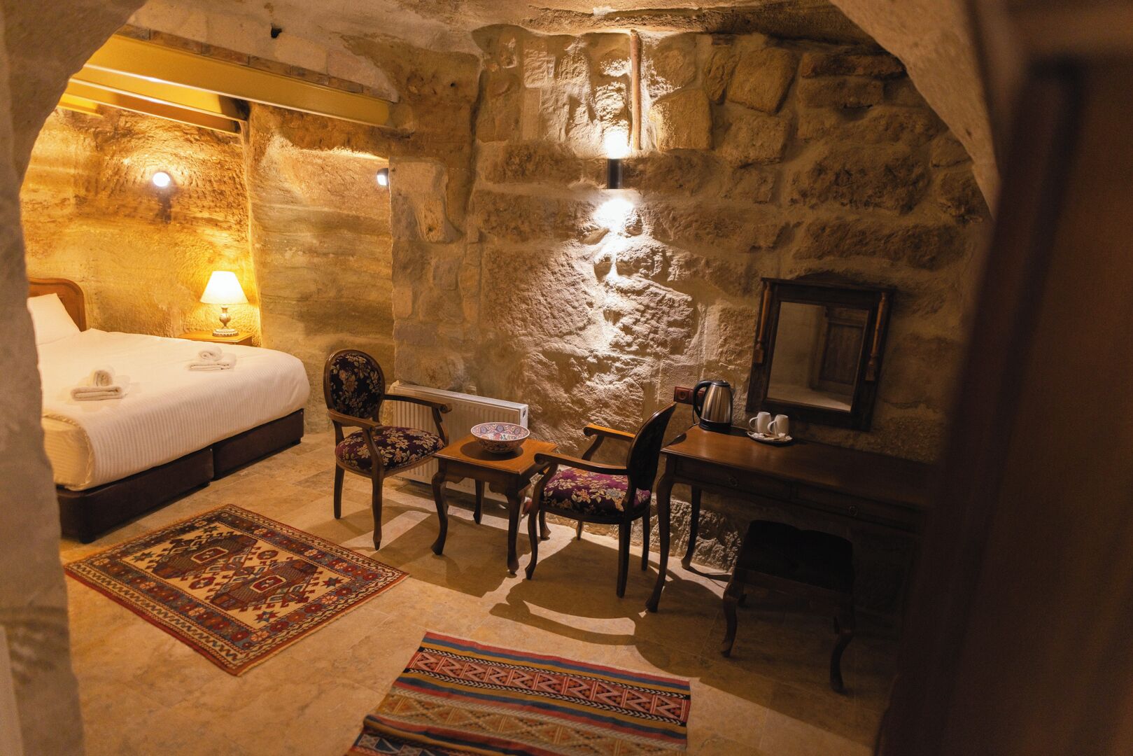 A cozy room with a stone wall, a bed, a chair, a small table with a bowl, and a radiator.