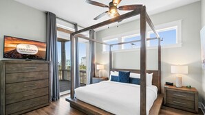 Un lit à baldaquin avec un cadre en bois, une télévision posée sur une commode et un balcon agrémenté de rideaux.