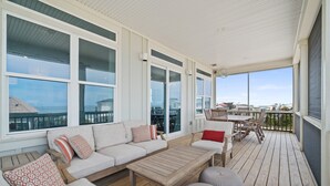 Une terrasse extérieure couverte, dotée d’une table et de chaises en bois, d’un canapé avec des coussins à motifs et de grandes fenêtres offrant une vue sur les environs.