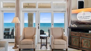 A living room with two beige armchairs, a small round table, a floor lamp, and a television on a wooden cabinet. Large windows offer a view of the ocean. - Turtle Tracks: Beachfront, East End, Pet Friendly, Sunset Beach (St. George Island)