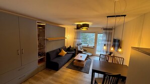 A modern living room with a sofa, coffee table, and dining table. There is a large cabinet with glass doors and a view of the outdoors through a window.
