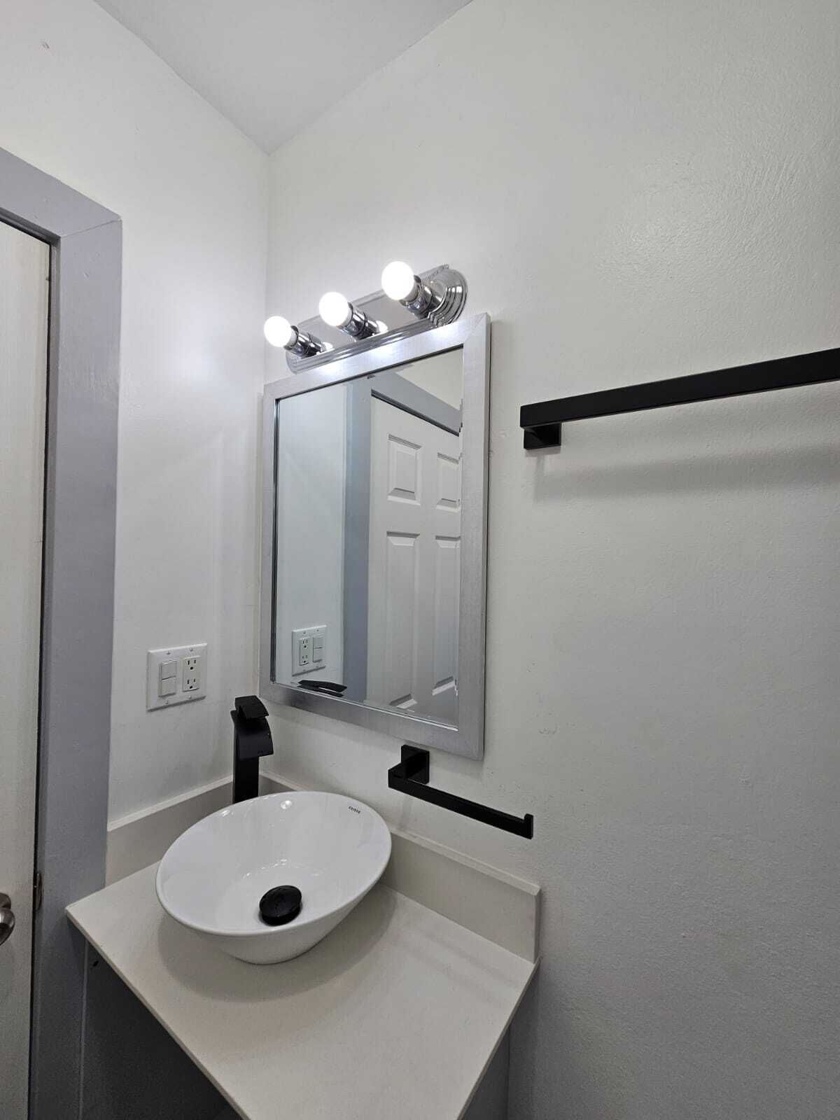 Casa de banho com lavatório branco, espelho com iluminação e porta-toalhas preto.