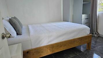 Un letto singolo con struttura in legno e biancheria bianca in una stanza con pavimento piastrellato scuro.