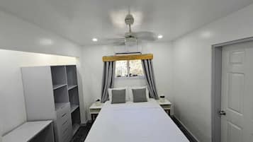 Une chambre à coucher comprenant un lit, deux tables de chevet, une armoire, un ventilateur de plafond et une fenêtre avec des rideaux.