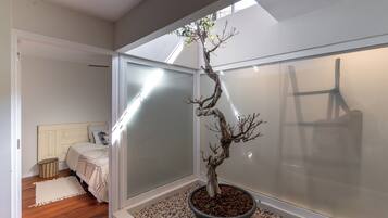 A room with a bed, a small stool, a potted bonsai tree, and a sliding glass door leading to another room.