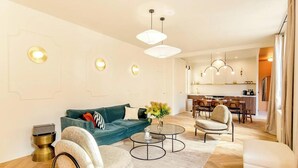 Una sala de estar moderna con un sofá verde, una mesa de centro redonda y un sillón beige. En el fondo, hay una zona de cocina con una mesa de comedor y sillas.