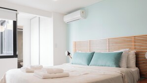 A hotel room with a bed, white linens, a window with blinds, an air conditioning unit, and a bedside table with a lamp. - MOT Santa Fe 5164 (Buenos Aires)