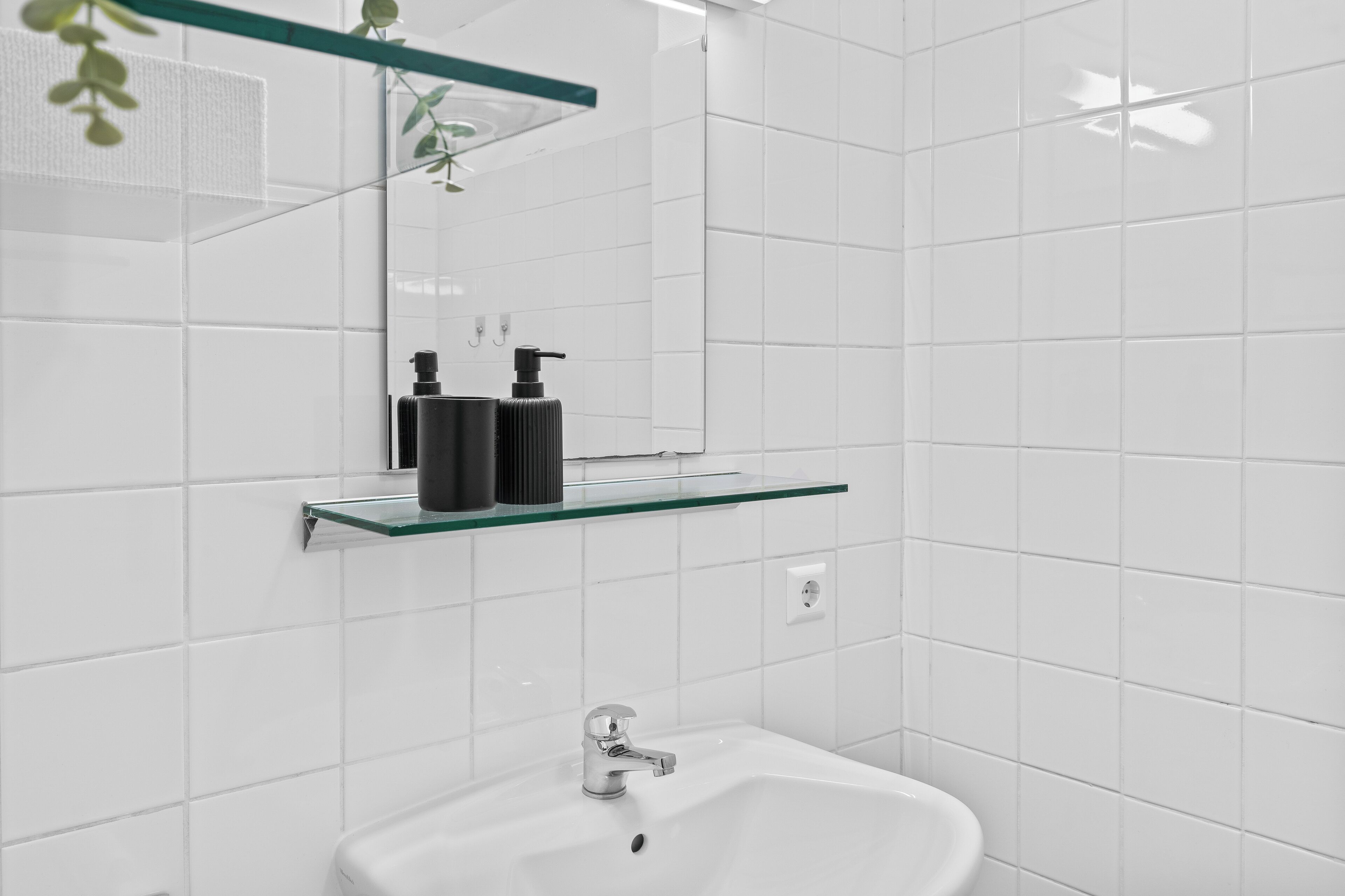 Un baño moderno con azulejos blancos, un estante de vidrio con artículos de tocador y un lavabo con grifo.