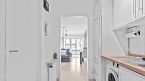 A modern kitchen with a built-in washer and dryer, a wooden countertop, and a view into a living room with a sofa and a window.