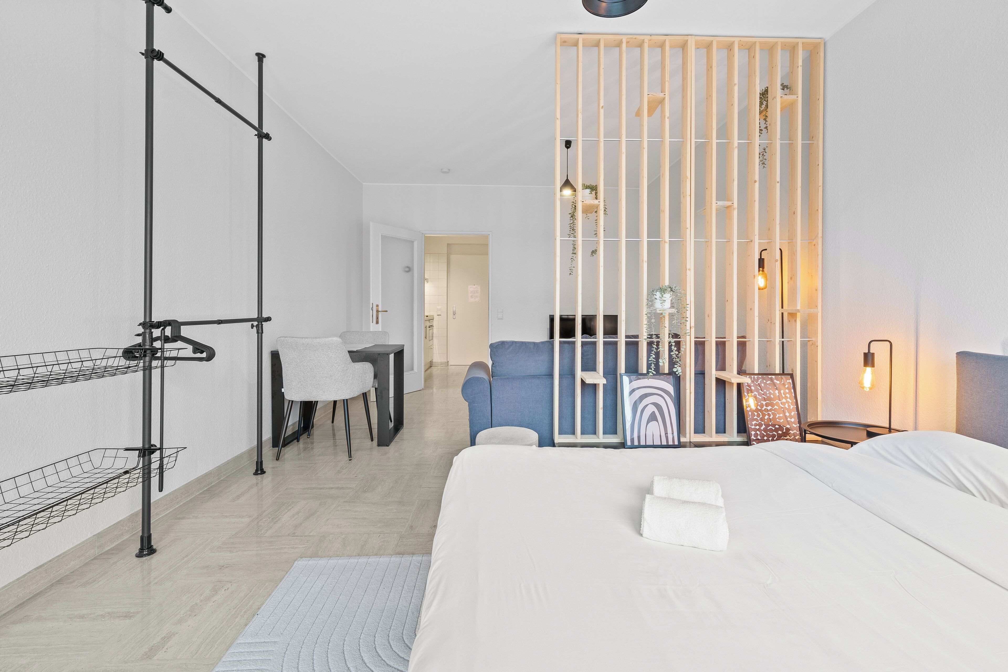 A modern hotel room with a large bed, a desk with a chair, a blue armchair, and a wooden partition with shelves.