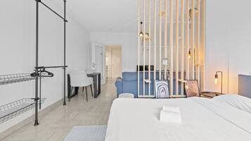 A modern hotel room with a large bed, a desk with a chair, a blue armchair, and a wooden partition with shelves.