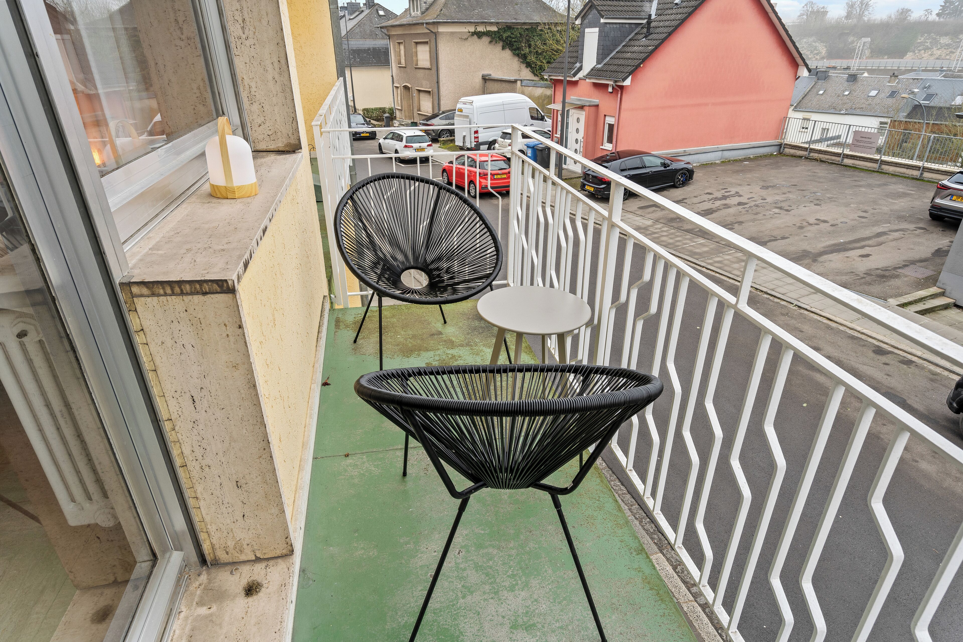 Ein Balkon mit zwei schwarzen Korbstühlen und einem kleinen Tisch, der auf einen Parkplatz mit mehreren Autos und einem Gebäude mit roter Fassade blickt.